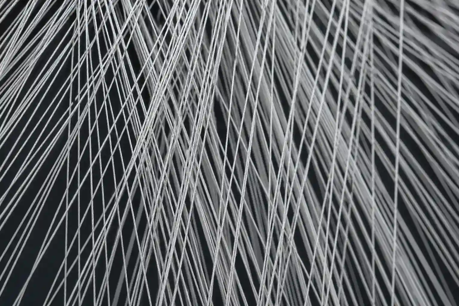Viele feine weiße und graue Fäden verlaufen diagonal und überkreuzen sich vor einem dunklen Hintergrund